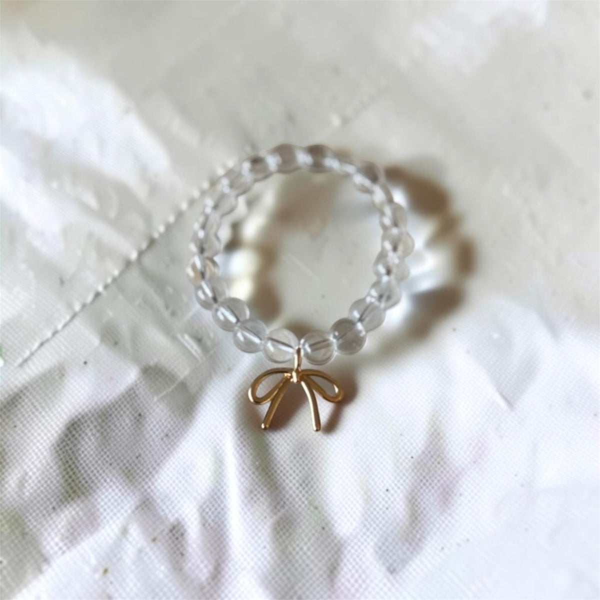 A beaded ring featuring clear, round glass beads and a small gold bow charm, resting on a textured white fabric background.