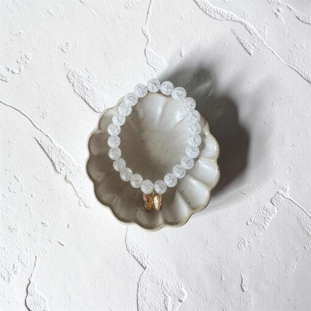 A beaded bracelet made of small clear faceted beads with a gold-toned connector, displayed on a scalloped ceramic dish against a textured white background.