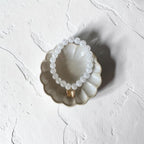 A beaded bracelet made of small clear faceted beads with a gold-toned connector, displayed on a scalloped ceramic dish against a textured white background.