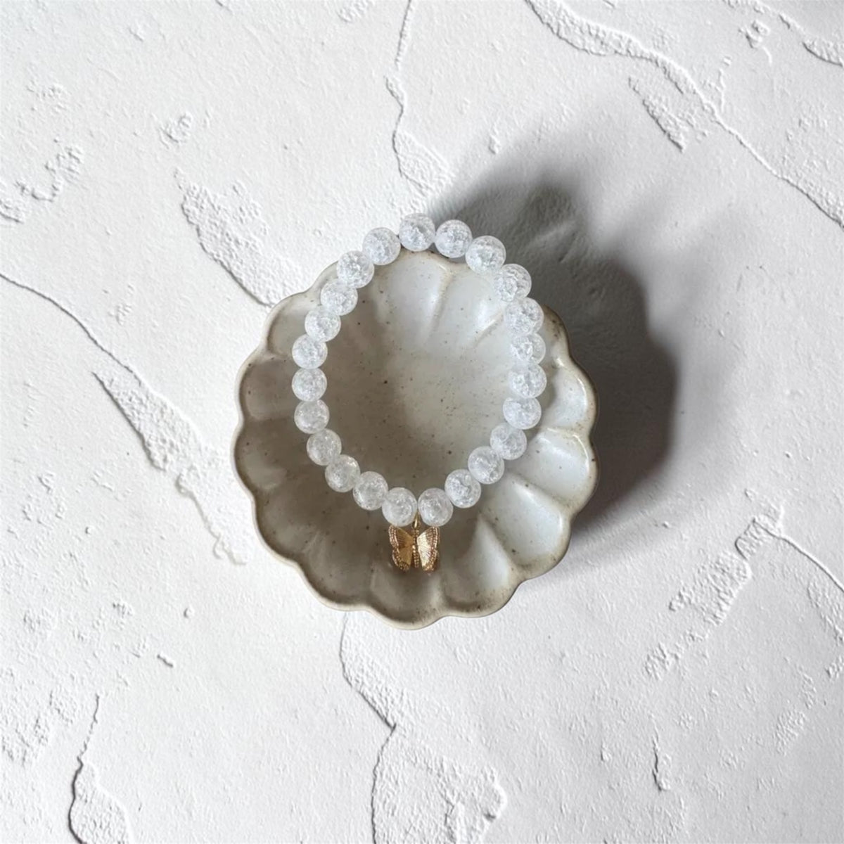A beaded bracelet made of small clear faceted beads with a gold-toned connector, displayed on a scalloped ceramic dish against a textured white background.