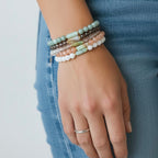 A close-up of a person's hand wearing a stack of five beaded stretch bracelets in pastel colors, paired with a silver band ring and blue jeans.