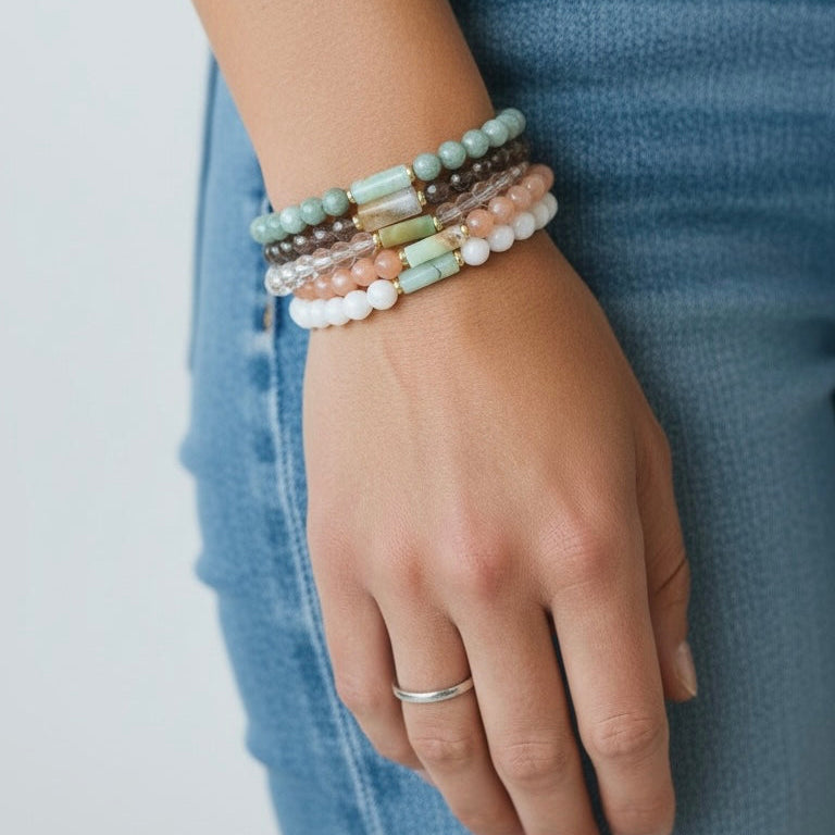A close-up of a person's hand wearing a stack of five beaded stretch bracelets in pastel colors, paired with a silver band ring and blue jeans.
