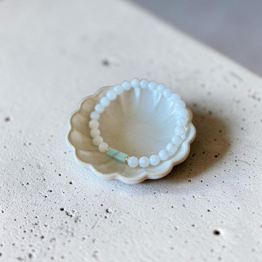 A delicate beaded bracelet with white round beads and a small mint green accent bead, displayed inside a small white ceramic dish on a textured concrete surface.