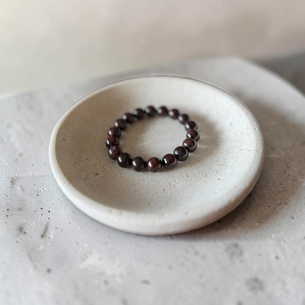 A beaded stretch bracelet made of dark brown polished round beads, displayed on a small round ceramic dish over a textured gray surface.