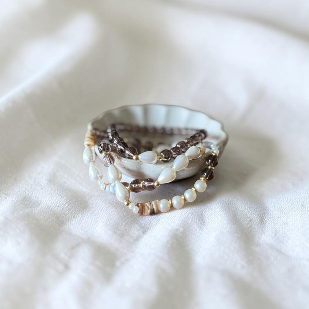 A stack of bohemian beaded bracelets in brown, cream, and white, displayed on a small scalloped dish over a soft white fabric background.
