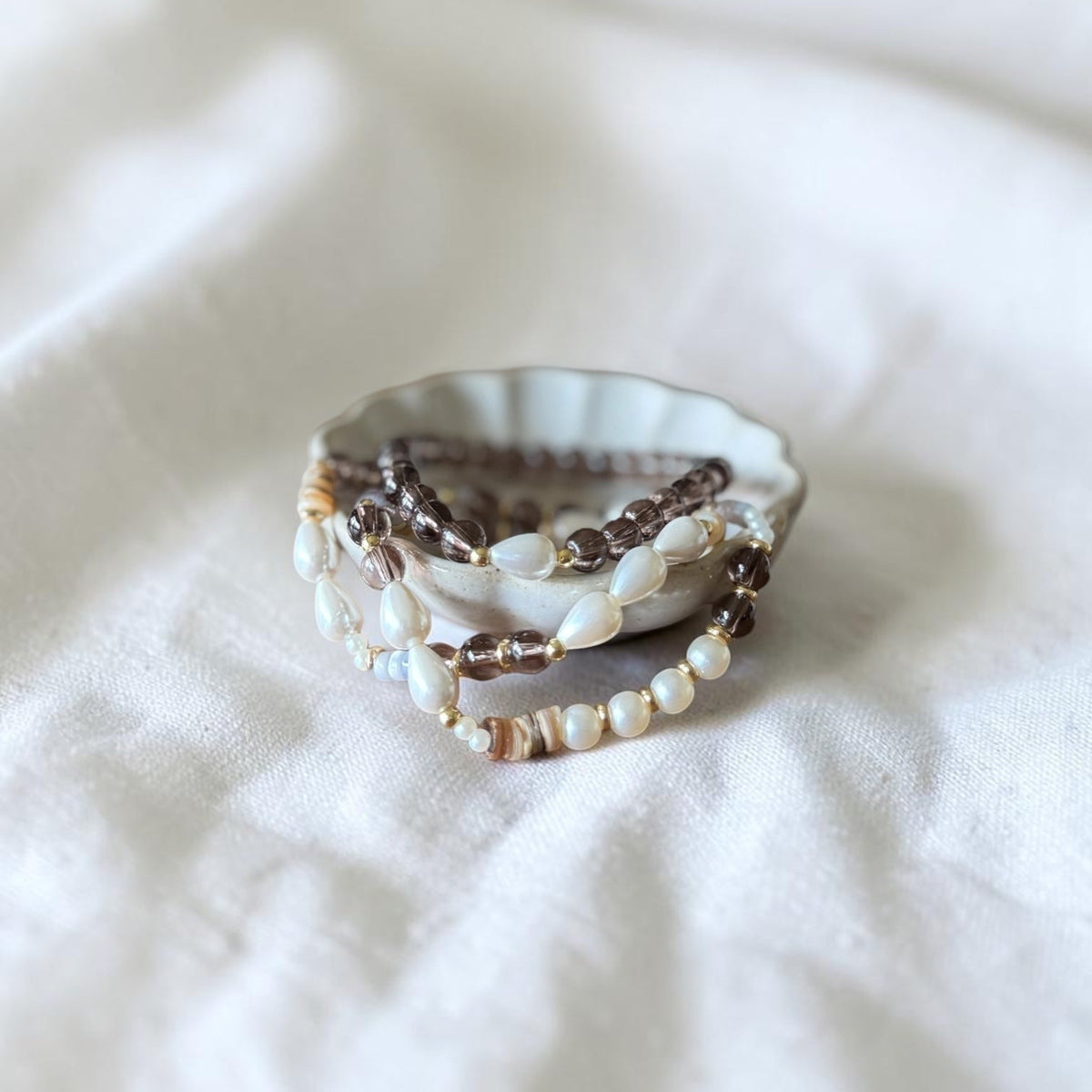 A stack of bohemian beaded bracelets in brown, cream, and white, displayed on a small scalloped dish over a soft white fabric background.