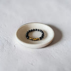 A beaded stretch bracelet with glossy black round beads and a textured gold-toned cylindrical accent, displayed on a small round off-white ceramic dish against a light gray textured background.