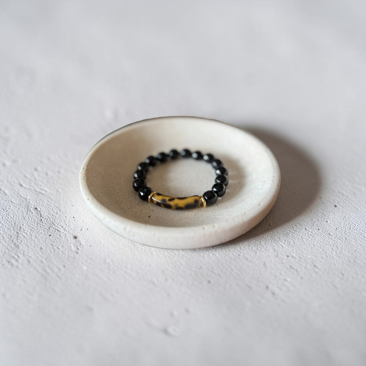 A beaded stretch bracelet with glossy black round beads and a textured gold-toned cylindrical accent, displayed on a small round off-white ceramic dish against a light gray textured background.