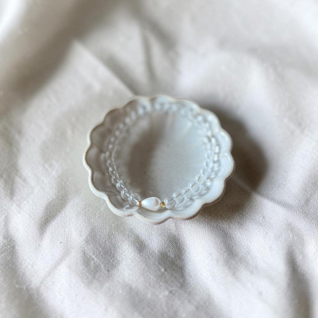 A delicate pearl beaded bracelet with a small gold accent, displayed inside a scalloped white ceramic dish on a textured white linen fabric background.
