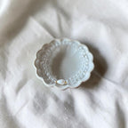 A delicate pearl beaded bracelet with a small gold accent, displayed inside a scalloped white ceramic dish on a textured white linen fabric background.