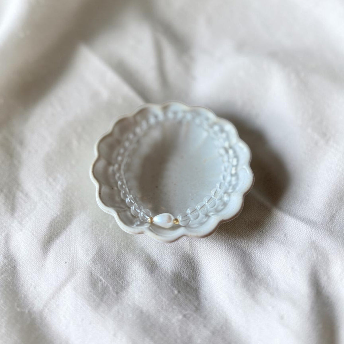 A delicate pearl beaded bracelet with a small gold accent, displayed inside a scalloped white ceramic dish on a textured white linen fabric background.
