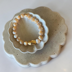 A beaded stretch bracelet with cream and white round beads and a single faceted gold accent bead, displayed on a small white ceramic dish over a larger speckled beige tray.