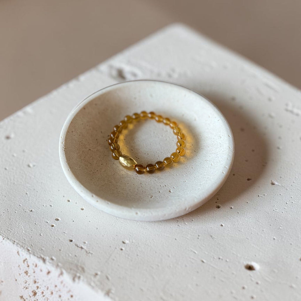 A small beaded stretch bracelet in a translucent amber-yellow color rests inside a shallow, round white ceramic dish, which is placed on a textured white stone block against a warm beige background.