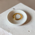 A small beaded stretch bracelet in a translucent amber-yellow color rests inside a shallow, round white ceramic dish, which is placed on a textured white stone block against a warm beige background.