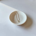 A delicate gold chain necklace with a small square pendant resting inside a small speckled ceramic dish on a white surface.