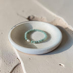A beaded stretch bracelet in light  green with a small white shell accent, displayed on a small round ceramic dish over a textured beige surface.