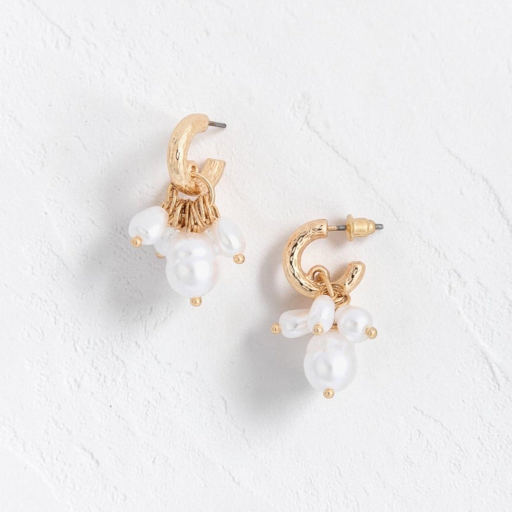 A pair of gold hoop earrings with dangling white pearl-like beads, displayed on a textured white surface.
