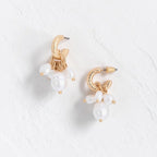 A pair of gold hoop earrings with dangling white pearl-like beads, displayed on a textured white surface.