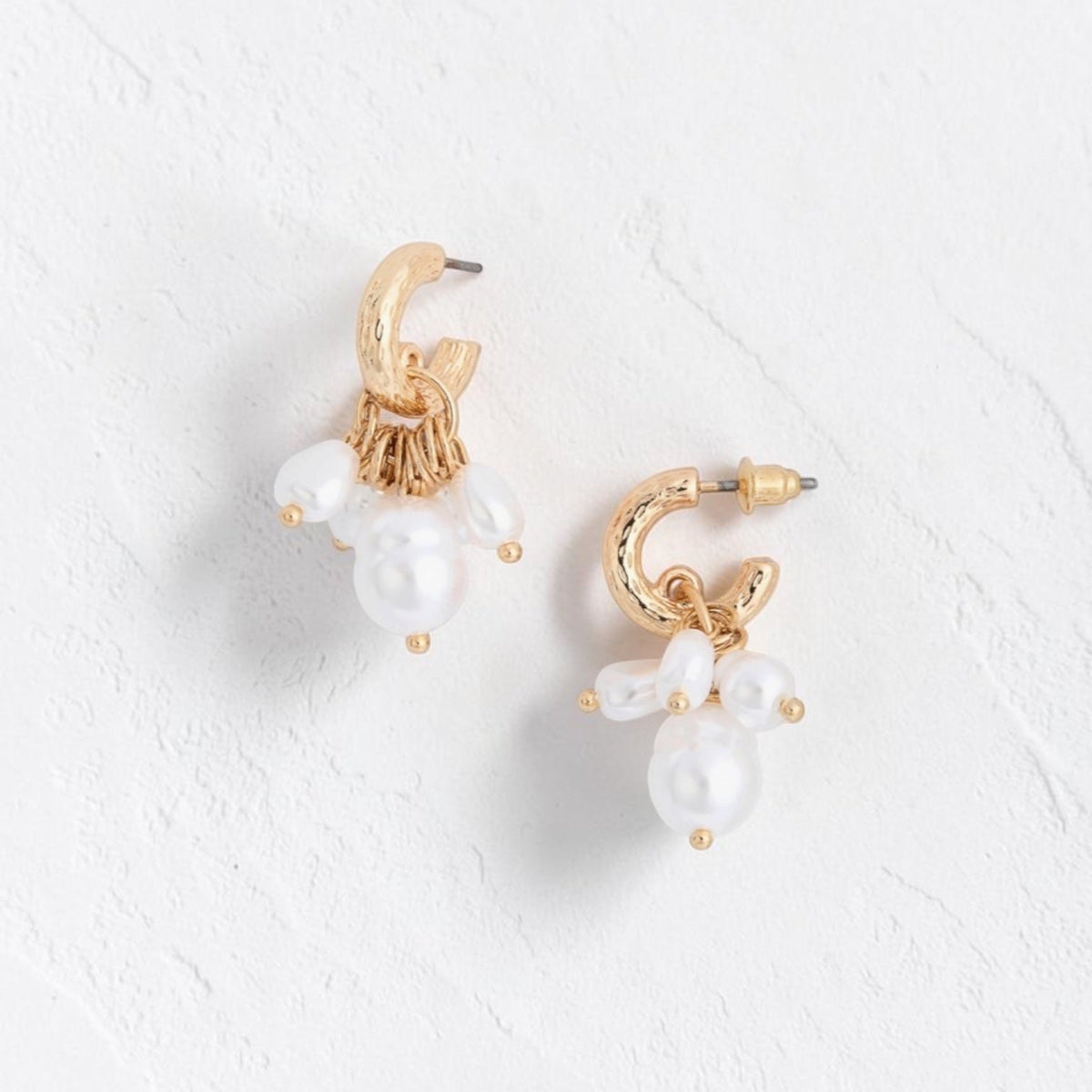 A pair of gold hoop earrings with dangling white pearl-like beads, displayed on a textured white surface.