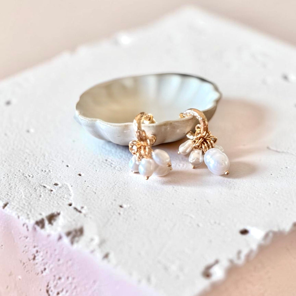 Gold hoop earrings with dangling white pearl-like beads, displayed on a small white dish atop a textured white surface against a soft beige background.
