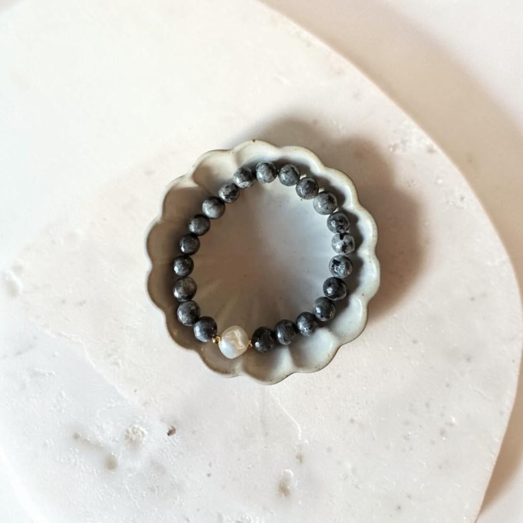 A beaded bracelet with dark gray to black round beads and a single white pearl accent, displayed inside a small scalloped ceramic dish on a light beige stone surface.