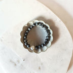 A beaded bracelet with dark gray to black round beads and a single white pearl accent, displayed inside a small scalloped ceramic dish on a light beige stone surface.