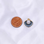 A small shell-shaped jewelry charm with a white pearl and blue enamel details, shown next to a copper penny for scale on a white fabric background.