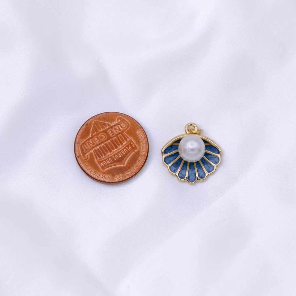 A small shell-shaped jewelry charm with a white pearl and blue enamel details, shown next to a copper penny for scale on a white fabric background.