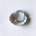 A beaded stretch bracelet in shades of blue and white, displayed inside a small scalloped ceramic dish on a textured white surface.
