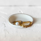 Two beaded stretch bracelets displayed in a small white ceramic dish. One bracelet features translucent amber-brown beads, while the other is made of similar amber beads with a single round white opal-like bead.
