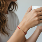 A close-up lifestyle shot of a woman wearing a delicate red and gold beaded bracelet on her wrist while holding a white ceramic mug.