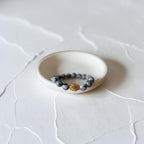 A beaded stretch bracelet with dark gray-blue round beads and a single faceted gold accent bead, displayed on a small white ceramic dish over a textured white surface.