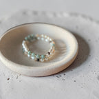 A delicate beaded stretch bracelet with light blue, white, and dark gray beads, displayed on a small round ceramic dish over a textured light gray surface.
