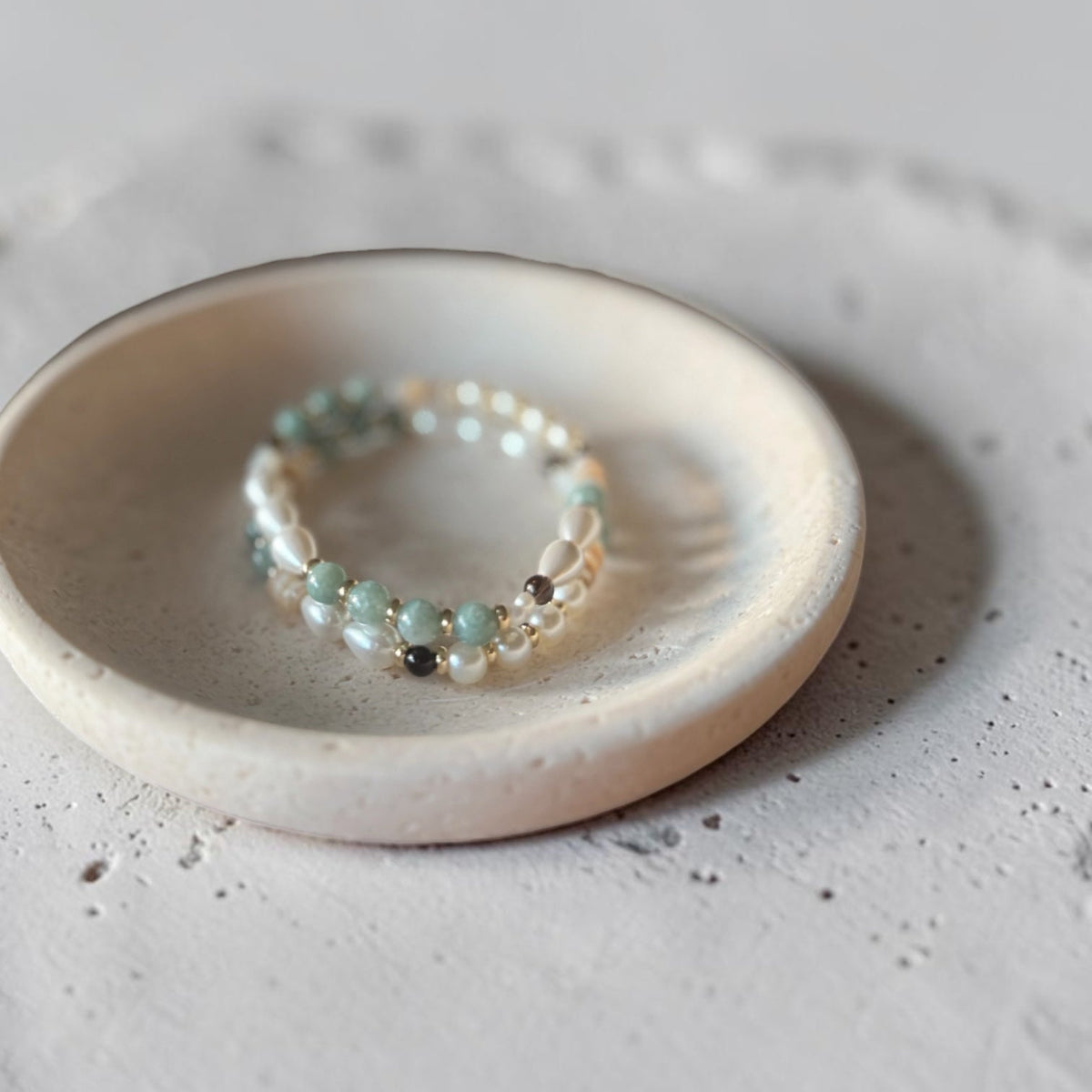 A delicate beaded stretch bracelet with light blue, white, and dark gray beads, displayed on a small round ceramic dish over a textured light gray surface.