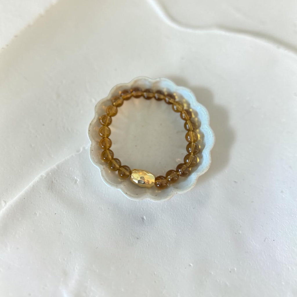 A beaded stretch bracelet with translucent amber-brown beads and a small faceted gold accent bead, displayed on a scalloped clear plastic stand against a white textured background.