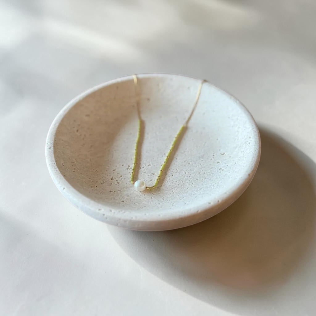 A delicate gold necklace with a small white bead pendant is displayed in a shallow, speckled off-white ceramic dish on a light surface.