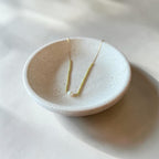 A delicate gold necklace with a small white bead pendant is displayed in a shallow, speckled off-white ceramic dish on a light surface.