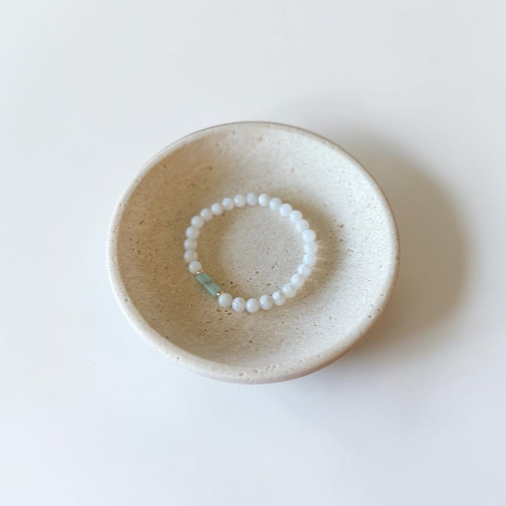 A beaded bracelet with white round beads and a small light blue accent bead, displayed inside a small speckled beige ceramic dish on a clean white background.