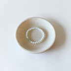 A beaded bracelet with white round beads and a small light blue accent bead, displayed inside a small speckled beige ceramic dish on a clean white background.