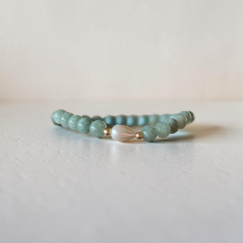 A beaded stretch bracelet featuring light teal-green round beads, a central white teardrop-shaped bead, and small gold spacer rings, resting on a light gray surface against a neutral beige background.