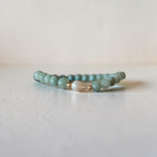 A beaded stretch bracelet featuring light teal-green round beads, a central white teardrop-shaped bead, and small gold spacer rings, resting on a light gray surface against a neutral beige background.