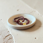 A beaded bracelet with dark red beads, a clear faceted stone, and a small gold heart charm rests on a small round ceramic dish, set against a textured beige concrete background.