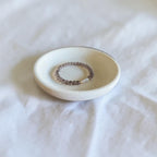 A beaded stretch ring with small brown beads and a white stone accent, displayed on a small round ceramic dish over a white fabric background.