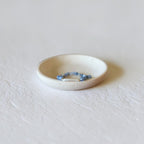 A small white ceramic dish holding a delicate beaded bracelet with blue and white beads, set against a textured white background.