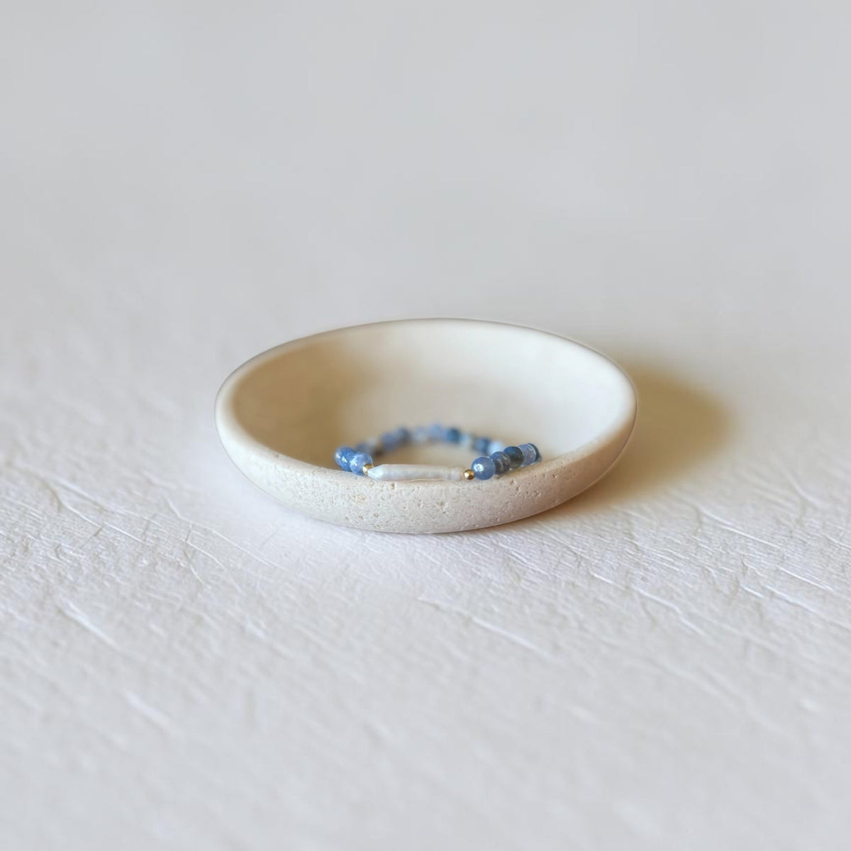 A small white ceramic dish holding a delicate beaded bracelet with blue and white beads, set against a textured white background.
