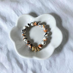A beaded stretch bracelet with white, black, and orange marbled beads and a small gold-toned bead accent, displayed on a white flower-shaped ceramic dish over a white fabric background.