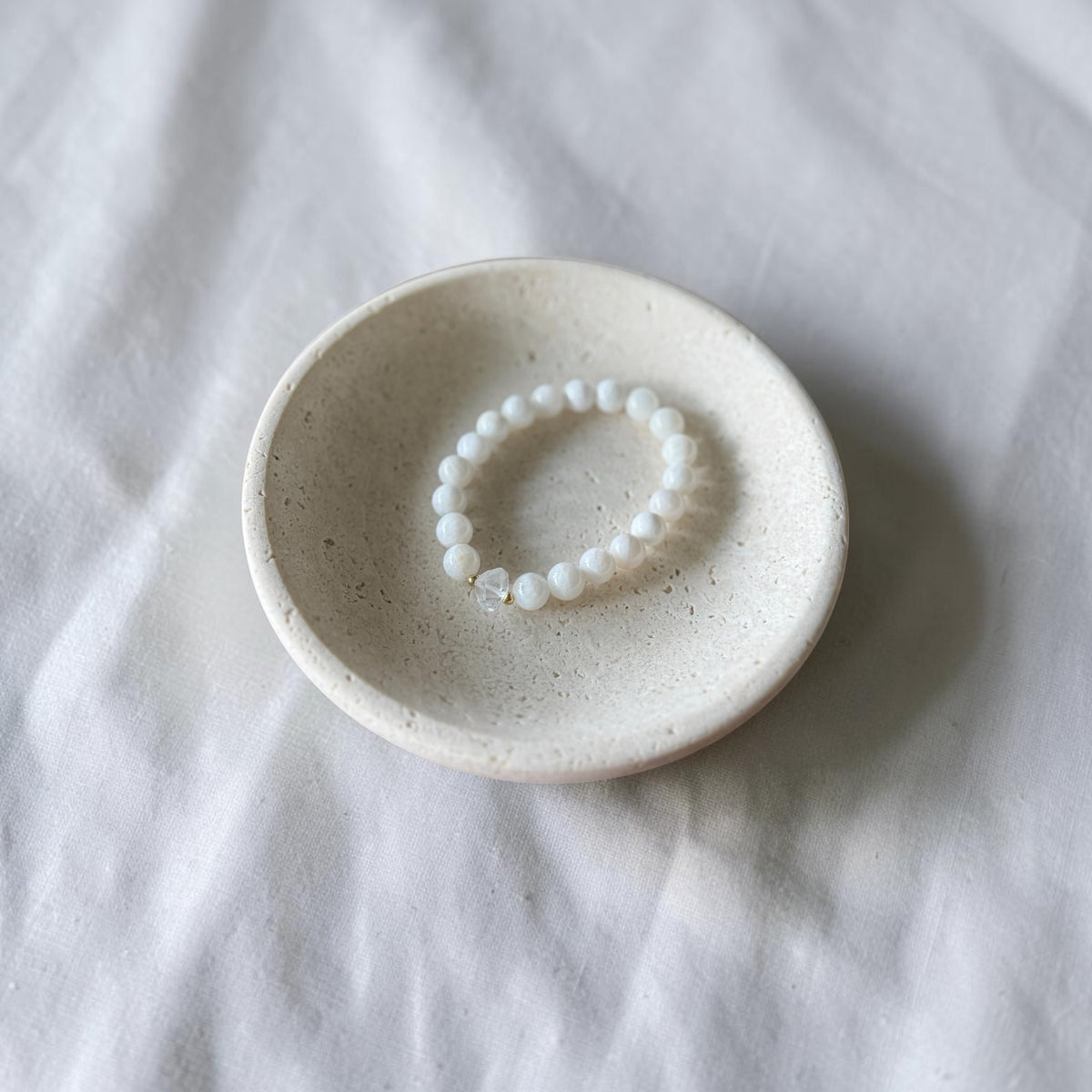 A white beaded bracelet with a small clear faceted bead, displayed inside a small speckled ceramic dish on a white fabric background.