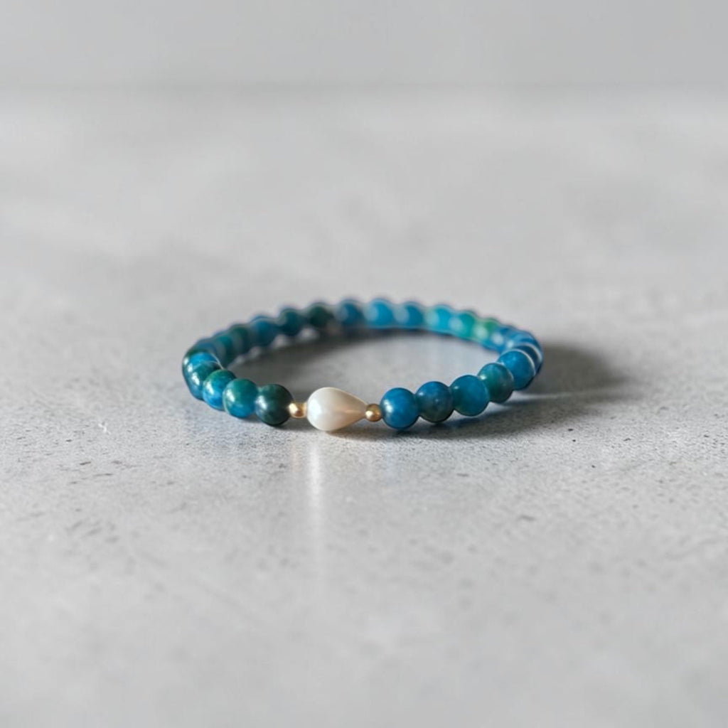 A beaded stretch bracelet with turquoise-blue beads and a single white teardrop bead, resting on a light gray textured surface.