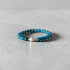 A beaded stretch bracelet with turquoise-blue beads and a single white teardrop bead, resting on a light gray textured surface.