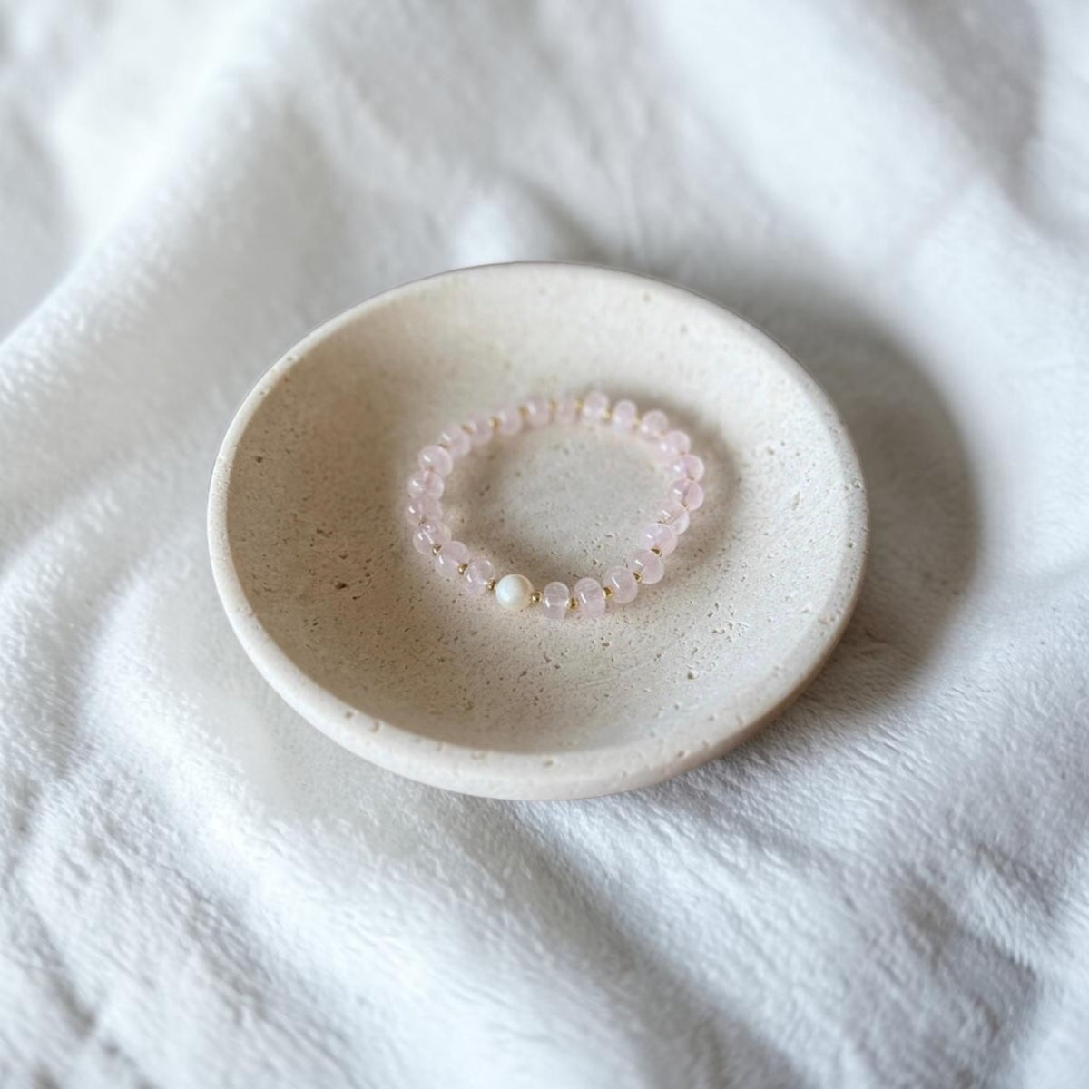 A delicate pink beaded bracelet with a single white pearl accent, displayed on a small speckled ceramic dish over a soft white textured fabric background.